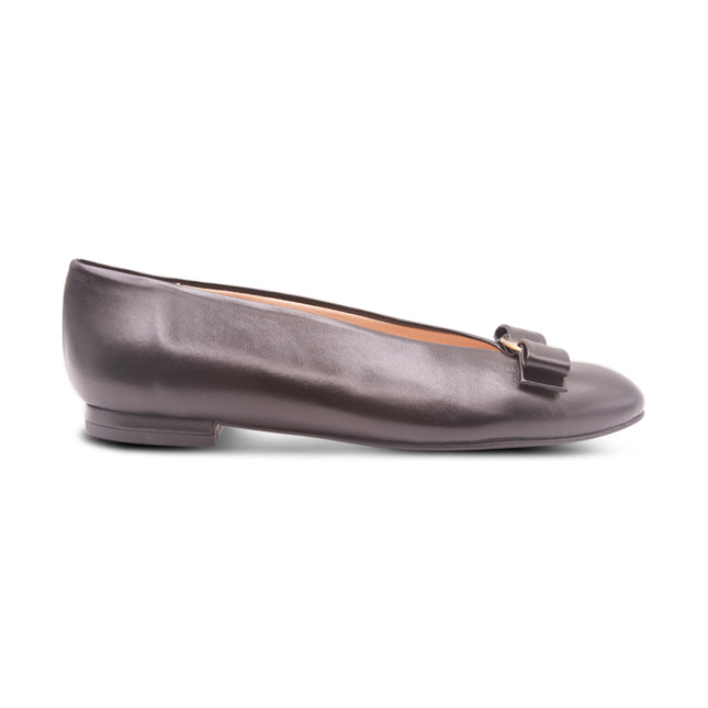 Black leather flat shown from the side, featuring a almond toe and a gold buckle detail with flatten bow across the front